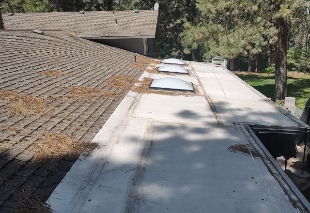 Before repair image showing a flat roof section with exposed underlayment next to a sloped, shingled roof area, surrounded by pine trees and a well-kept yard in Spokane