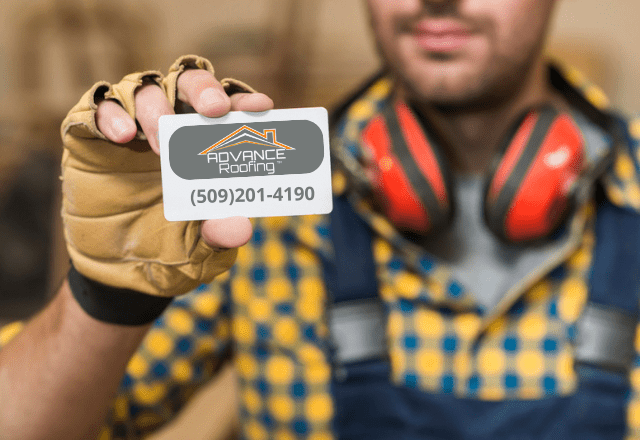 A worker in protective gloves and earmuffs holds up a business card for Advance Roofing LLC, with the contact number (509)201-4190 clearly displayed