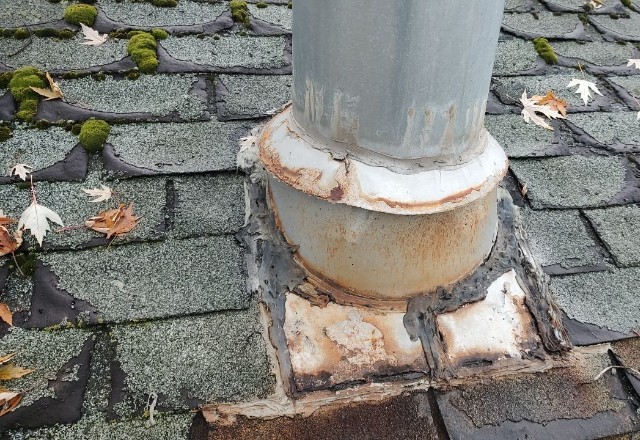Roof vent pipe with deteriorated and rusty flashing on an asphalt shingle roof, requiring urgent repair to prevent leaks