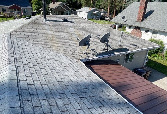 View of an aged asphalt shingle roof in Spokane Valley with satellite dishes installed and lush greenery in the background.
