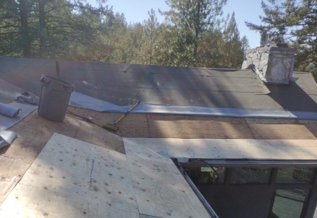 Partial view of a roof under repair in Spokane Valley, showing exposed underlayment and a stray hammer, with construction debris and natural backdrop