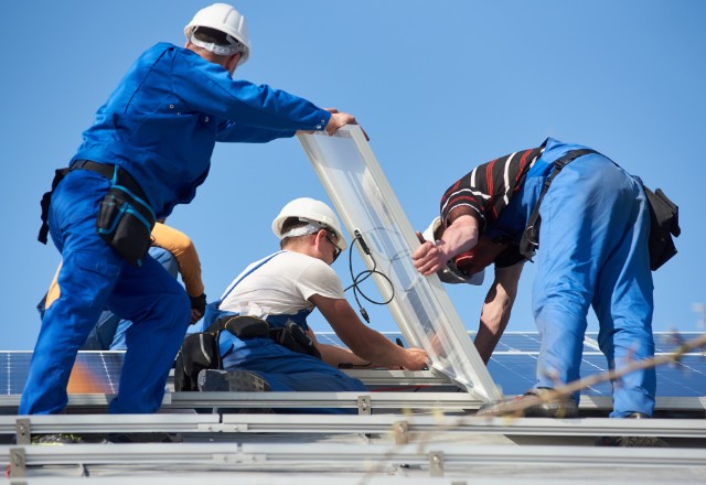 A team of Advance Roofing LLC workers in blue uniforms and safety helmets installing a new window on a roof with solar panels in Spokane, indicating professional window replacement services with a focus on energy efficiency.