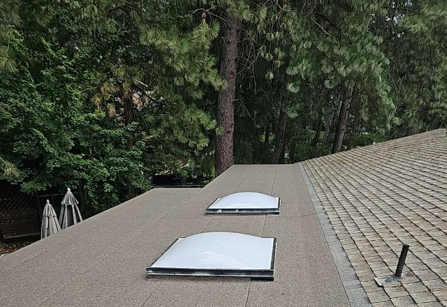 After repair image depicting a flat roof with new skylights installed, adjacent to an older shingle roof, overlooking tall pine trees in Spokane