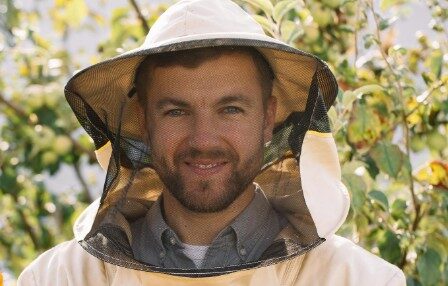A man with a smile, wearing a protective suit and hat with a veil, demonstrating protection against stinging insects.