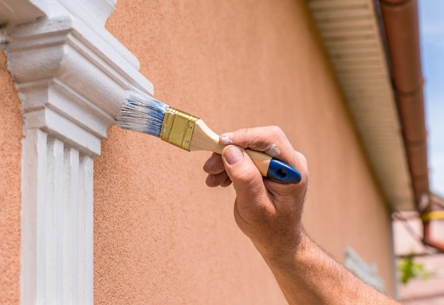 Close-up of a person hand applying white paint to a detailed column exterior with a brush, against a backdrop of a peach-colored wall and brown gutter in Spokane