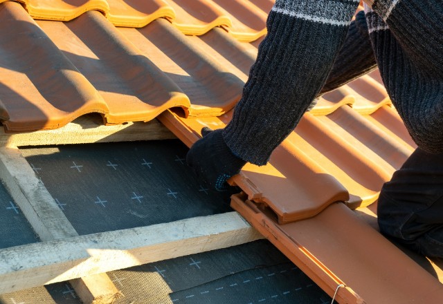 Close-up of hands installing terracotta tiles on a roof, with a focus on the precise placement of the tiles over a black waterproof underlay and wooden roof battens.