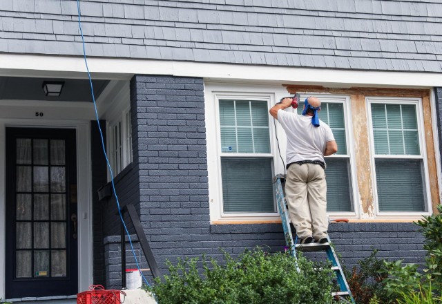Residential painter on a ladder painting window trims on a gray house, representing dedicated residential painting services available in Spokane