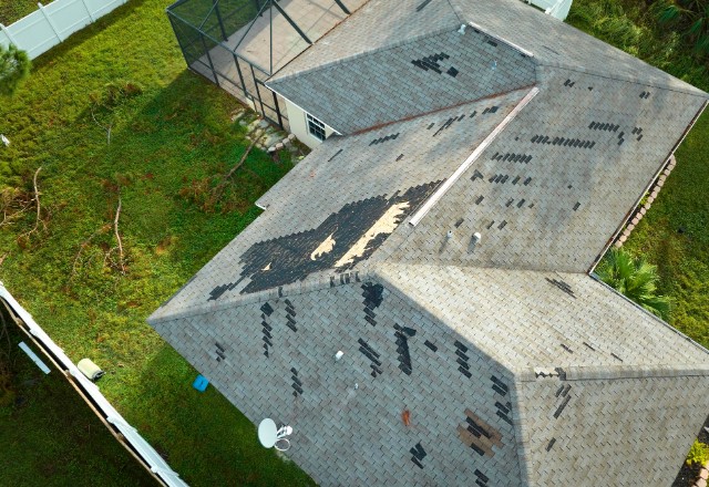 The image depicts a residential house roof that has sustained significant damage, likely due to storm winds. There are numerous shingles missing, exposing the underlying roofing material.