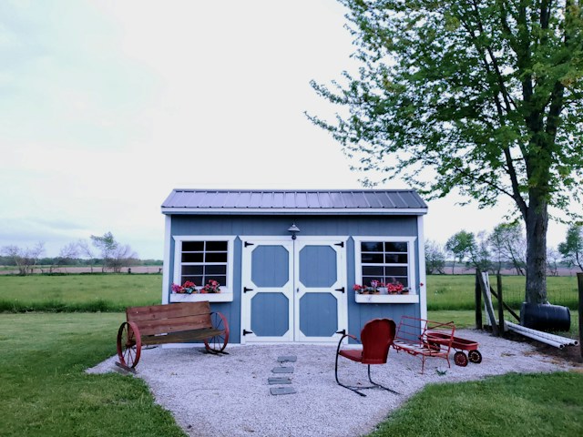 A charming garden shed, freshly repaired with a gray metal roof and sky blue doors, complemented by decorative red accents and planters, situated in a lush green yard.
