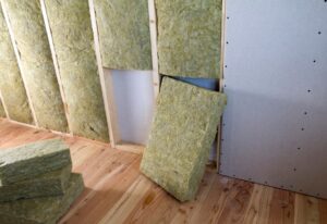 Interior view of an attic under construction with rockwool insulation batts being installed between wooden studs, beside unfinished drywall, for enhanced energy efficiency.