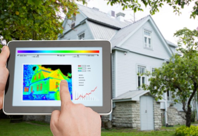 A person's hand points to an infrared image on a tablet, showcasing different temperatures across the roof of a house in the background, indicative of an infrared roof inspection. This diagnostic approach highlights areas of heat loss or insulation issues in the residential building