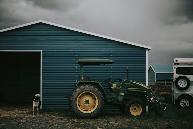 A modern blue metal shed with a clean, sturdy roof, a parked tractor to the side, and a dog standing guard, set against a dramatic cloudy sky.