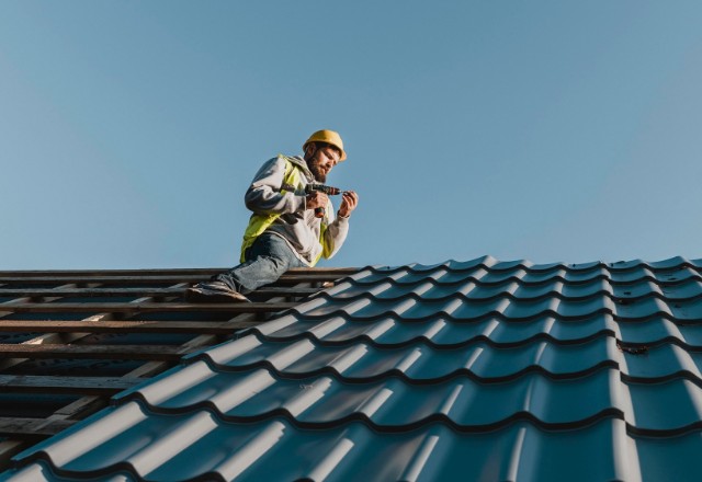A roofer wearing a yellow hard hat and safety vest is installing blue tiles on a steep roof, representing skilled tile roofers in Spokane under a clear sky