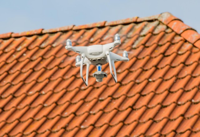 A drone equipped with a camera hovers in front of a terracotta-tiled roof, performing a drone-assisted roof inspection. The image captures the modern approach to roofing assessments, with a clear sky in the backdrop emphasizing the inspection activity