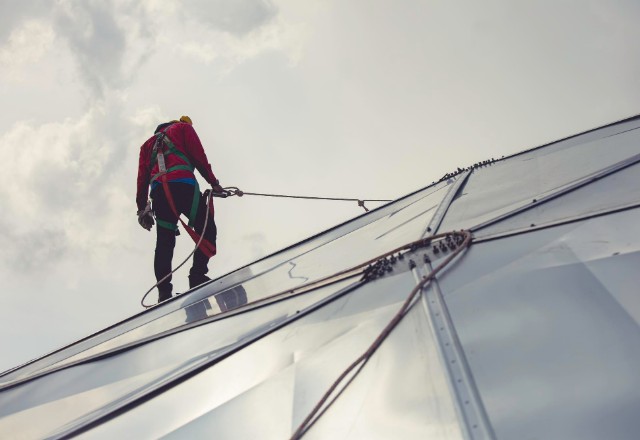 A licensed contractor performing professional roof cleaning, securely harnessed and using rope access techniques on a steep, reflective metal roof, demonstrating specialized roofing services against a cloudy sky backdrop.