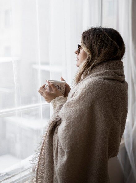 A woman wrapped in a cozy beige blanket stands by a window, holding a mug, suggesting a chilly atmosphere possibly due to poor insulation, a common issue in Spokane households seeking energy efficiency.