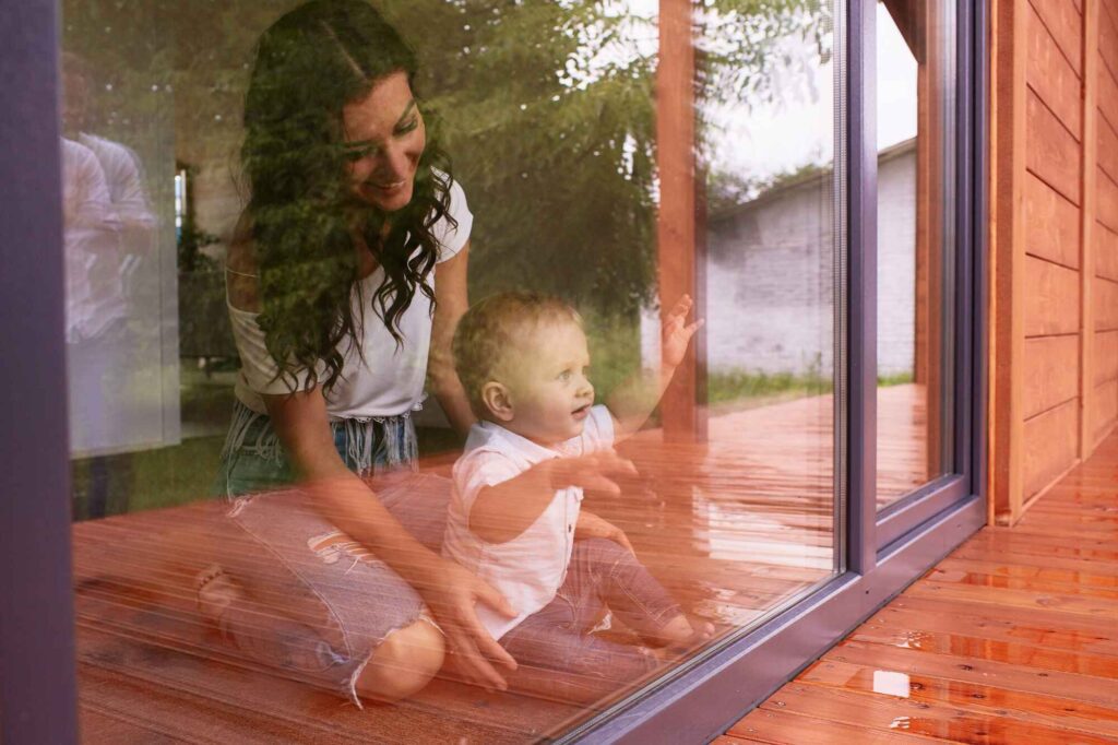 A mother and her young child enjoy a playful moment by a large window in Spokane, WA, with the reflection showing a serene backyard, highlighting the quality installation by a local window contractor