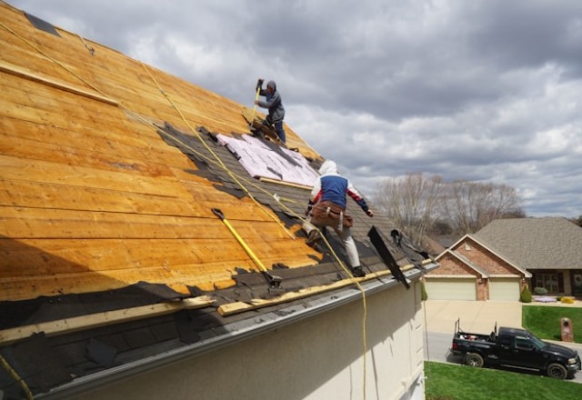 Workers engaged in a full roof replacement process, shown installing underlayment on wooden sheathing, with various roofing tools in use on a residential home, against a backdrop of a cloudy sky and suburban neighborhood.