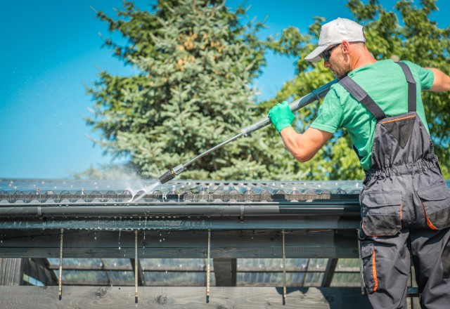 Worker in green shirt and white cap uses a high-pressure washer to clean a black rain gutter on a roof, typical for rain gutter cleaning services in Spokane.