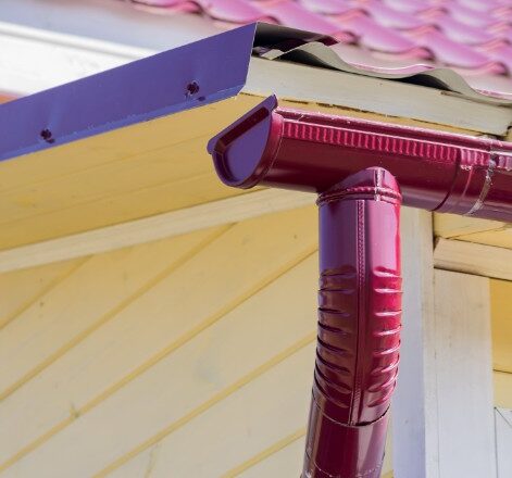 A vivid red gutter and downspout against a purple trim and yellow siding, highlighting the vibrant color options for gutters in Spokane homes