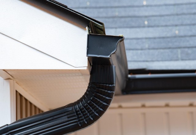 A close-up of a black gutter downpipe connecting to the gutter system under the white eave of a house, exemplifying popular gutter styles in Spokane