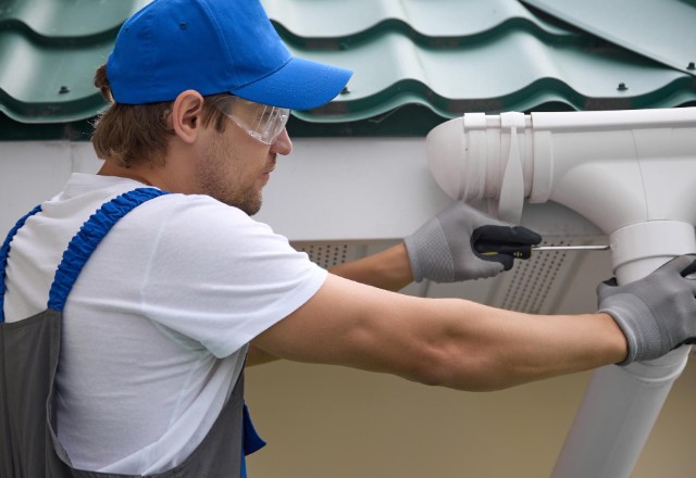 A skilled installer in a blue cap and safety goggles is fastening a new white rain gutter on a green metal-tiled roof, representing the installation of rain gutters in Spokane.