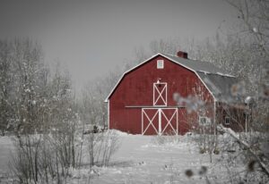 Rustic red pole barn nestled in a tranquil winter forest, crafted by skilled pole barn builders in Spokane, WA