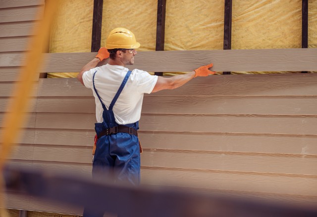 Construction worker in blue overalls and yellow hard hat carefully reattaching beige siding to a house exterior, demonstrating how to repair siding that fell off.