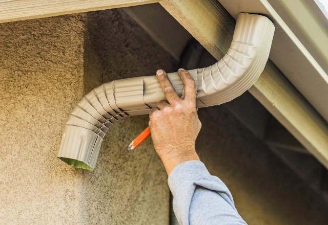 A worker's hand is shown attaching a beige aluminum downspout to a fascia under the roof, a process commonly seen in gutter installation in Spokane