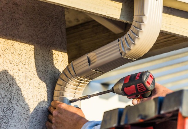 Hand of a worker securing a curved beige gutter downspout to a house wall with a red cordless drill, illustrating a typical gutter repair service in Spokane