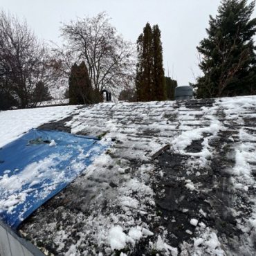 A roof with patches of snow and a blue tarp weighted down by snow, indicating a temporary fix for a potential leak or damage