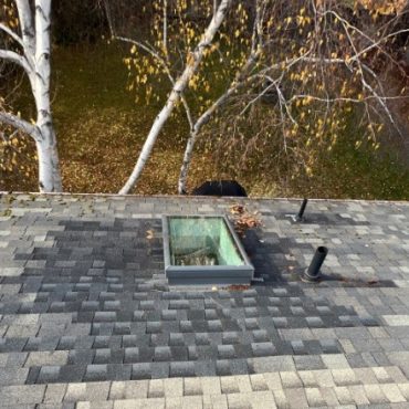 Elevated view of a residential asphalt shingle roof featuring a skylight, with surrounding trees in autumn, suggesting a serene neighborhood setting